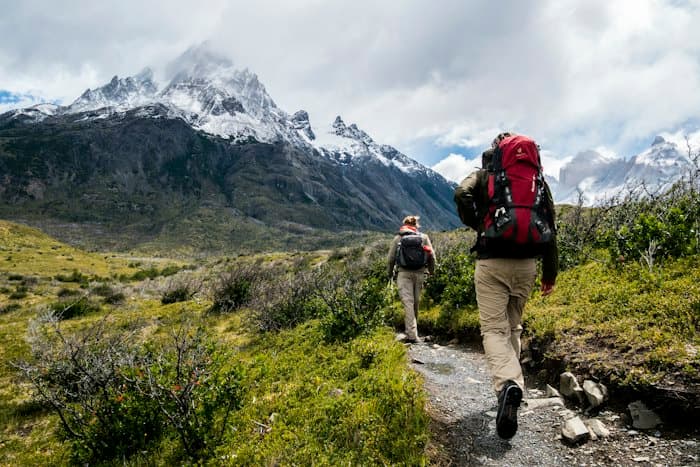 Two hikers with backpacks walking a trail towards snow-capped mountains